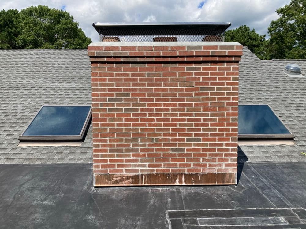 Brick chimney on a roof with skylights and a cloudy sky backdrop.