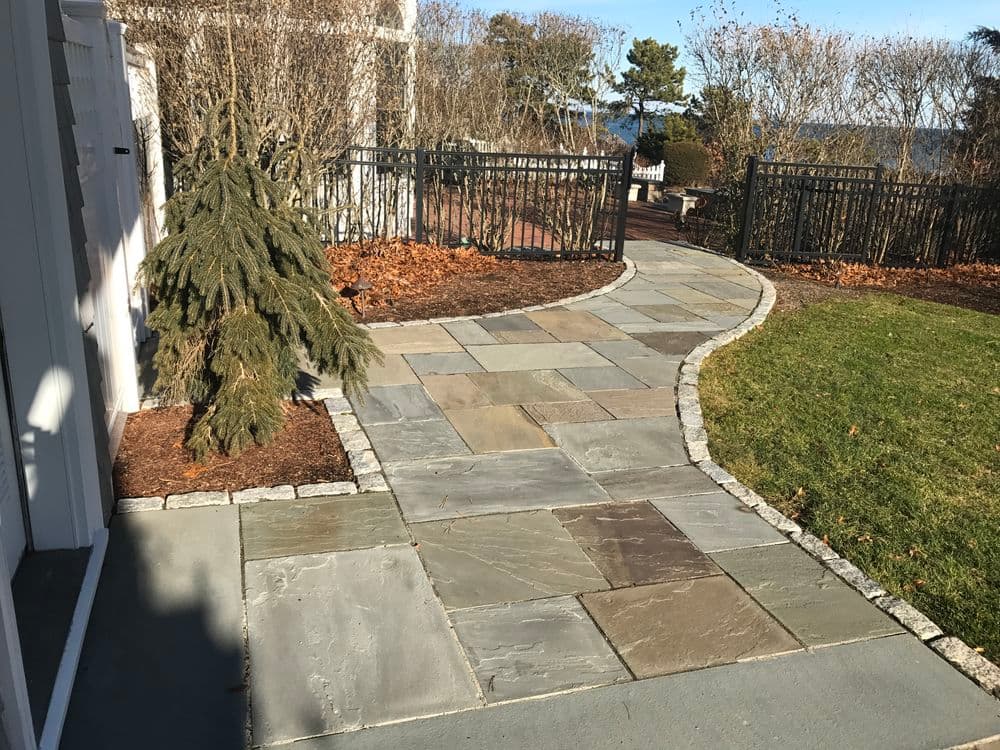 Curved stone walkway with greenery and a fence in a scenic outdoor setting.