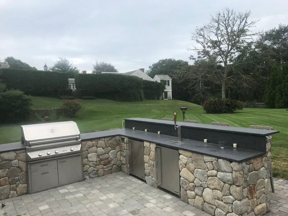 Outdoor kitchen with stone countertop, grilling station, and lush green landscaping.