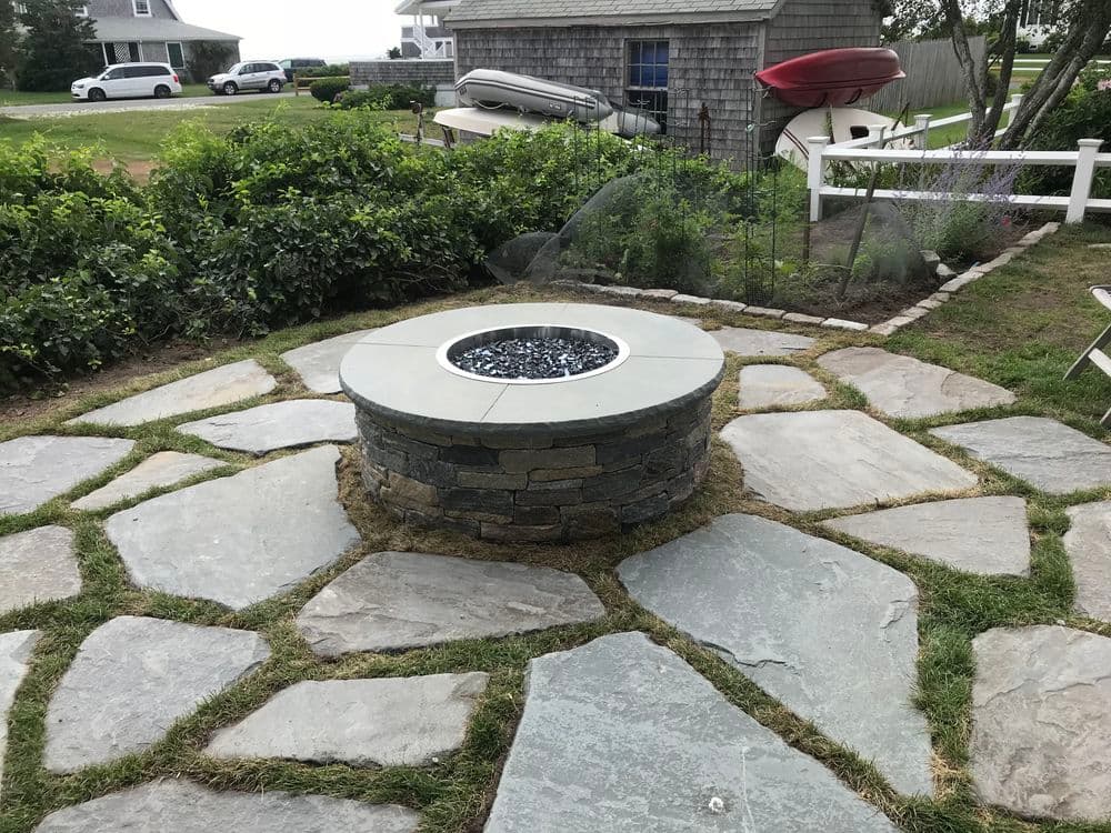 Stone fire pit surrounded by a flagstone patio in a landscaped outdoor area.