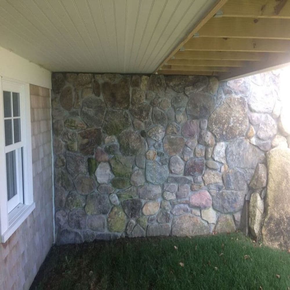 Stone wall with various textures and colors beneath a wooden porch, surrounded by grass.