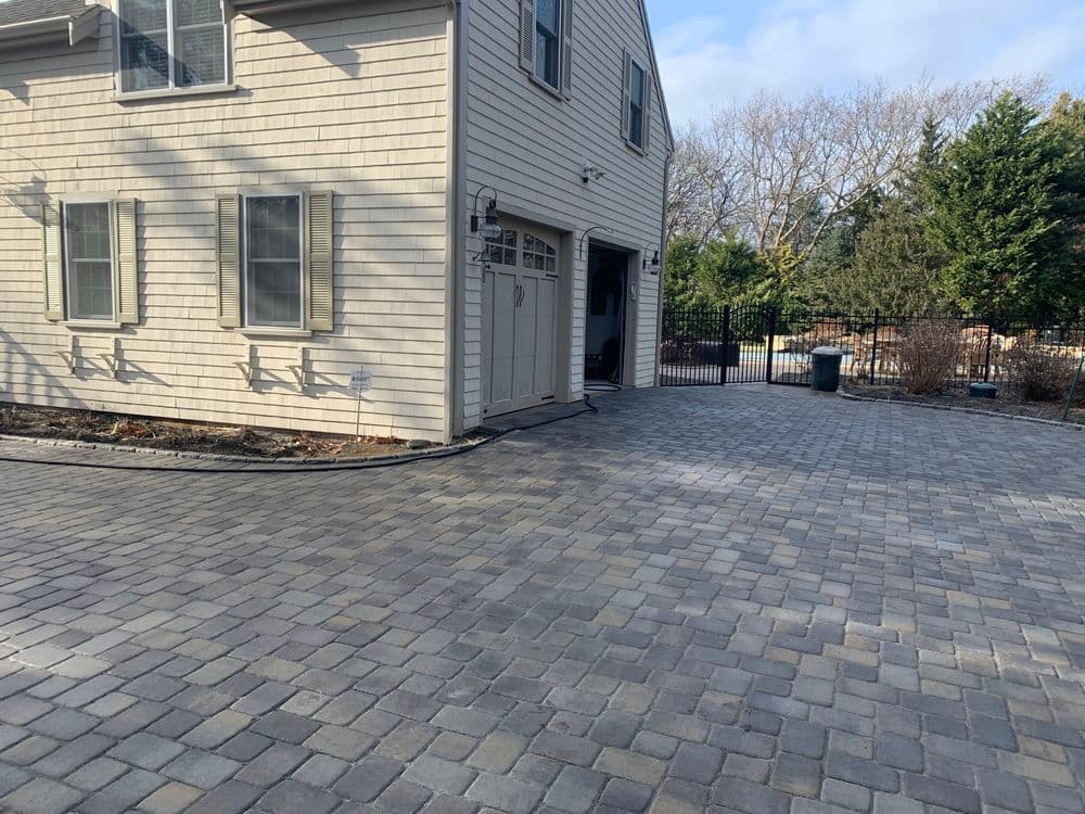 Paved driveway beside a house with stone pattern, surrounded by greenery and a fence.