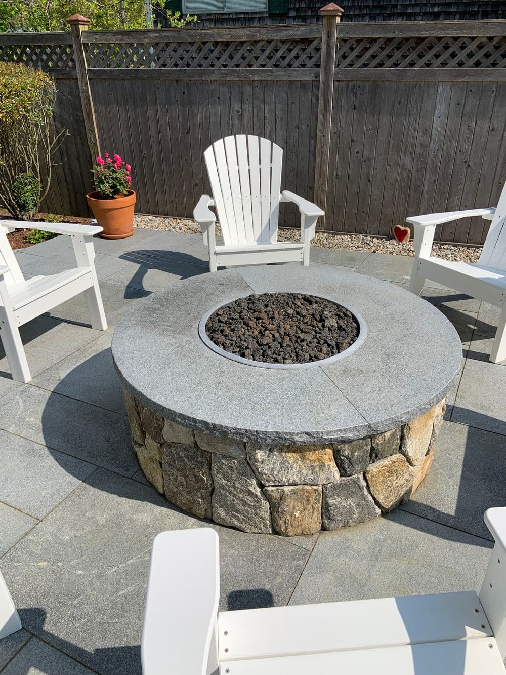 Stone fire pit surrounded by white Adirondack chairs in a landscaped backyard.
