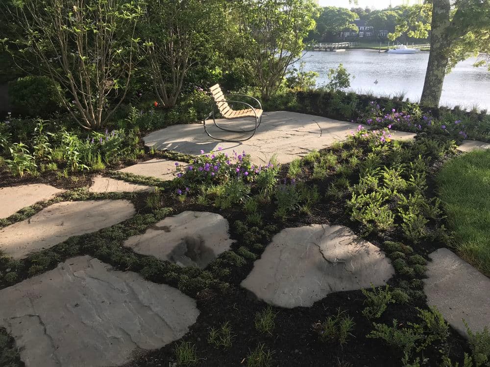 Lakeside garden with stone pathway, purple flowers, and a lounge chair nestled among greenery.