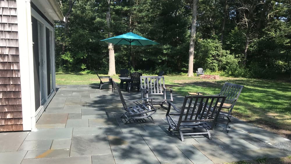Patio area with black chairs, a table, and a blue umbrella surrounded by trees.