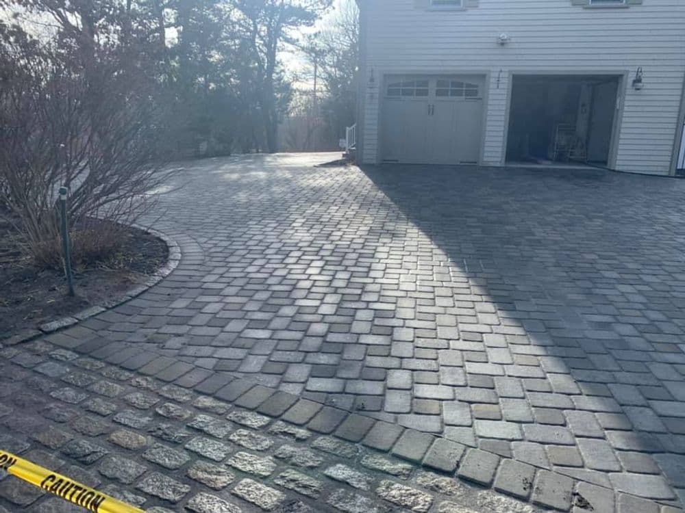 Paved driveway with cobblestone design and caution tape, leading to a house in sunlight.