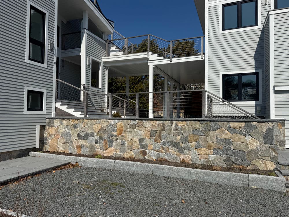 Modern home exterior with stone wall, deck, and stylish railing against a clear blue sky.