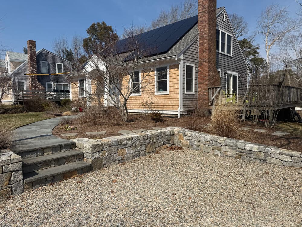 Charming coastal home with solar panels, stone walkway, and landscaped yard in bright daylight.